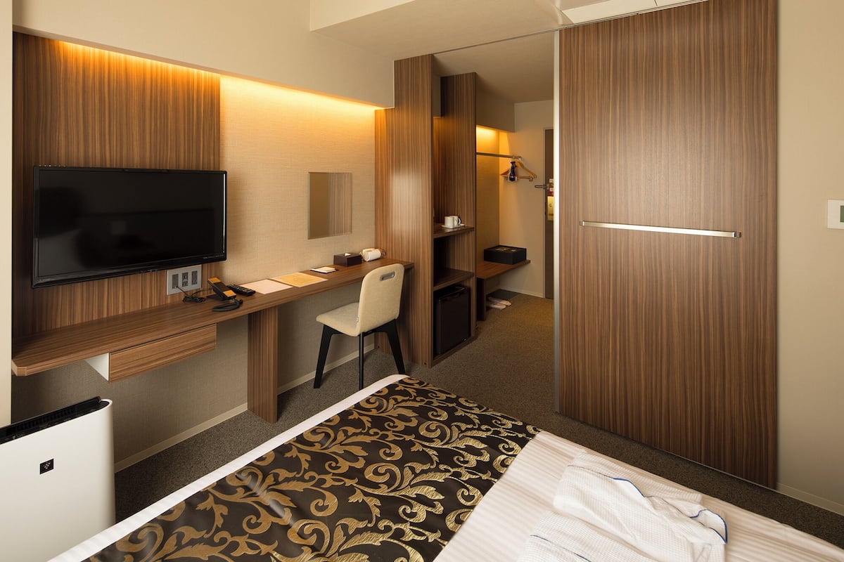 A hotel room with a bed, a desk with a flat-screen TV, a chair, a wardrobe, and a mirror.