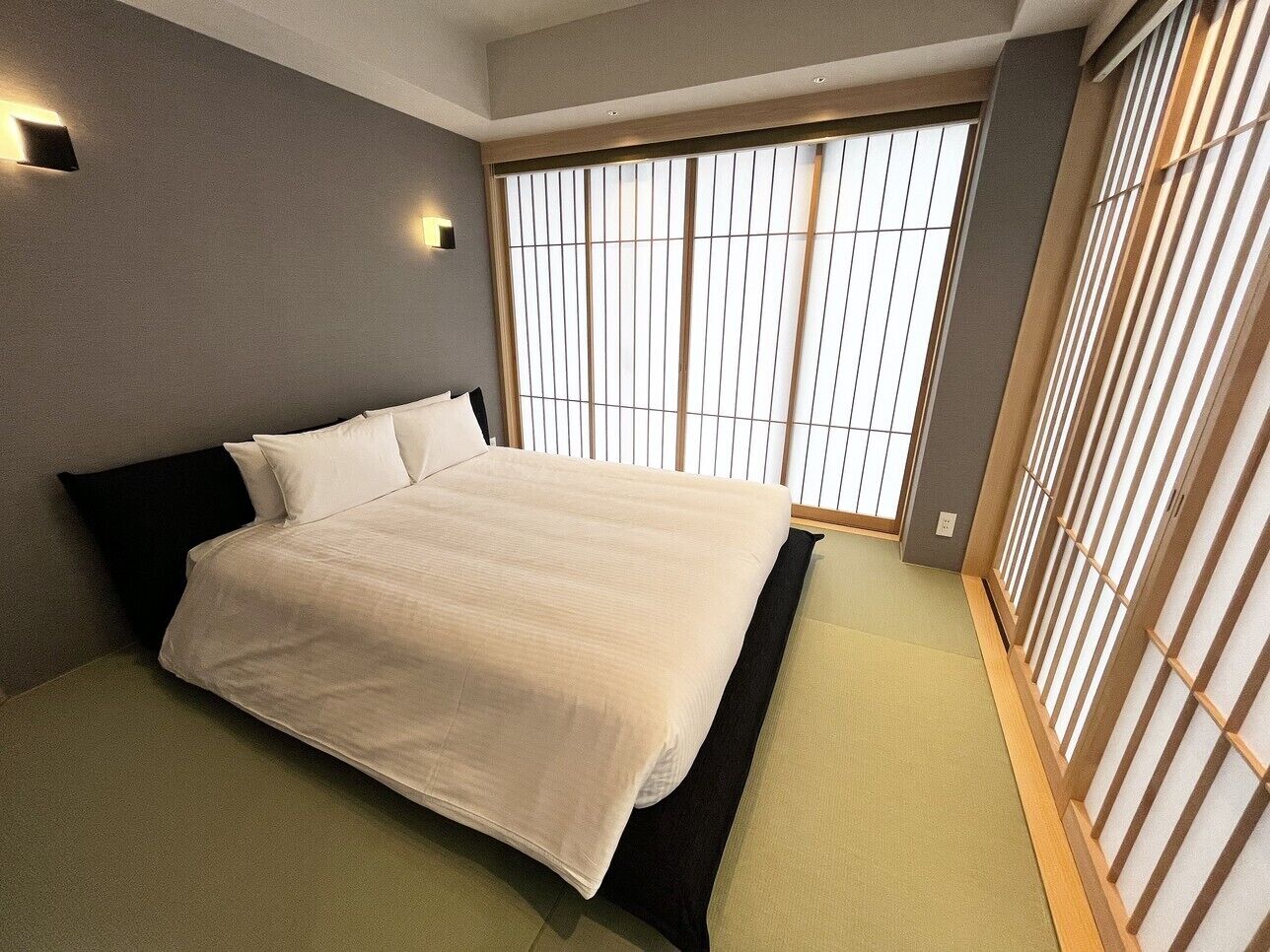 A bedroom with a bed, a window with blinds, and a wall-mounted light.