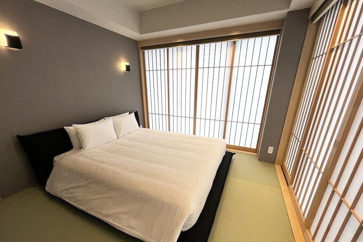 A bedroom with a bed, a window with blinds, and a wall-mounted light.