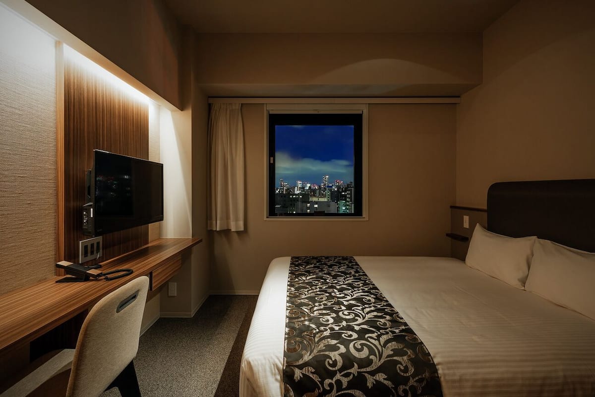A hotel room with a bed, a desk with a television, a chair, and a window with a city view.