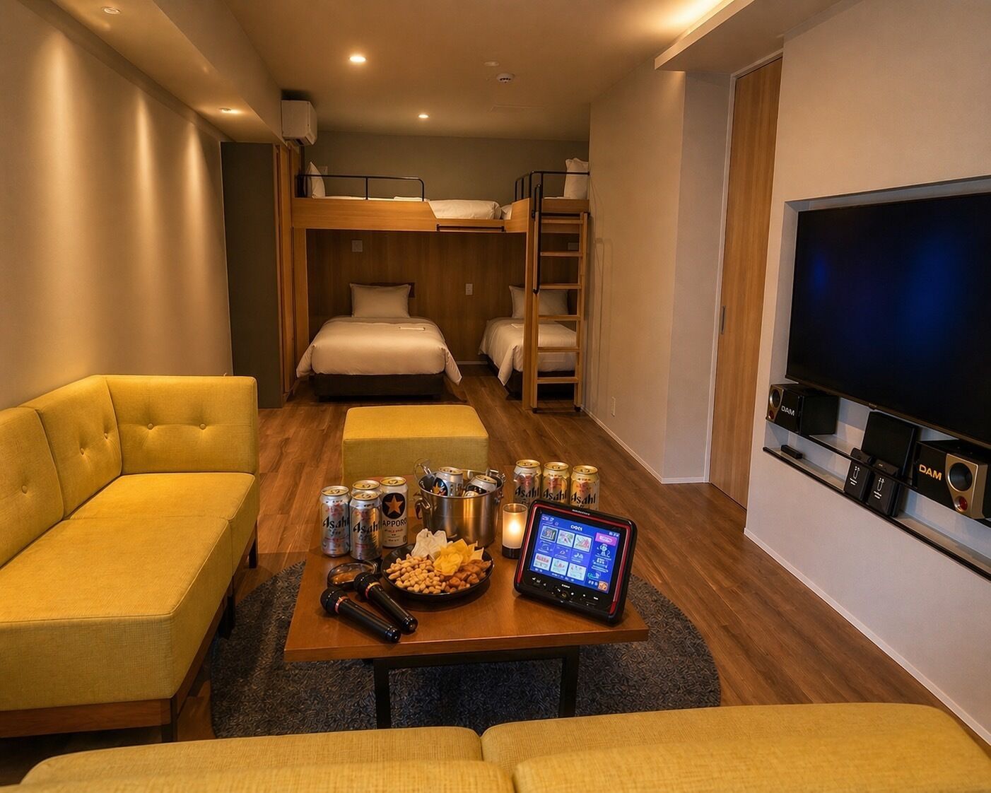 A hotel room with a sofa, bunk beds, a TV, a table with snacks and drinks, and a digital device.