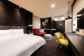 Quarto de hotel com uma cama grande, um sofá vermelho, uma cadeira amarela, uma mesinha redonda e uma escrivaninha com telefone e controle remoto.