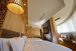 Quarto de hotel moderno com uma cama grande, cabeceira de madeira, televisão e área de estar com sofá e cortinas.