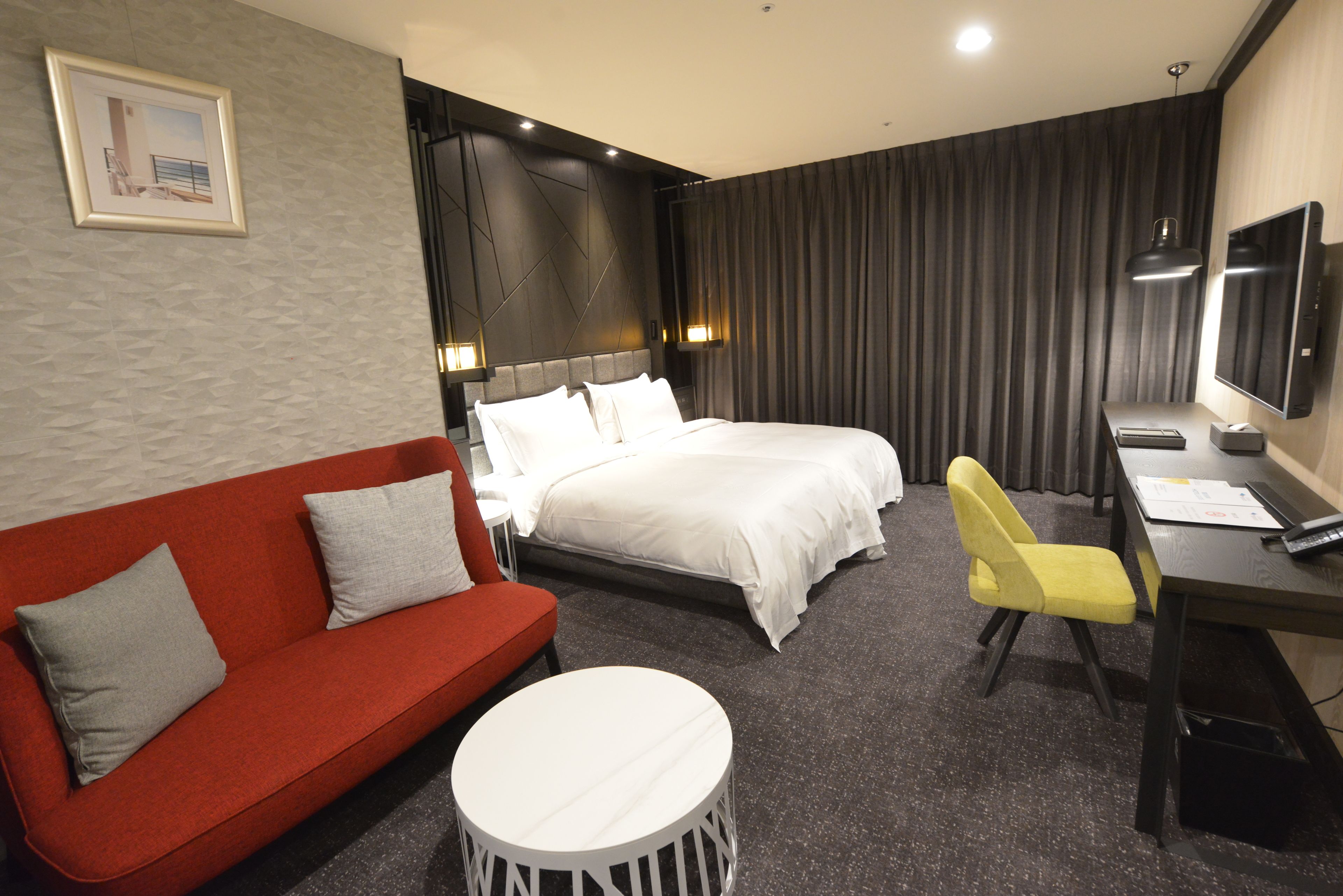 A hotel room with a red sofa, two beds, a desk with a chair, a round white table, and a TV.
