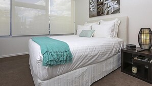 Un dormitorio con cama, mesita de noche, lámpara y ventana con persianas.