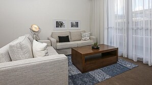 Una sala de estar moderna con un sofá, una mesa de centro y una lámpara. Hay cuadros enmarcados en la pared y un ventanal con cortinas translúcidas.