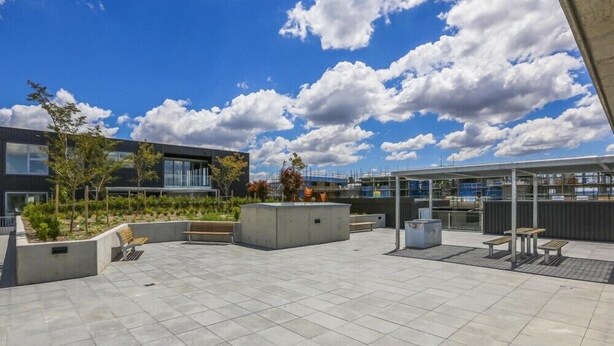 Una terraza en la azotea con un edificio moderno, zonas de estar y un cielo despejado con nubes.