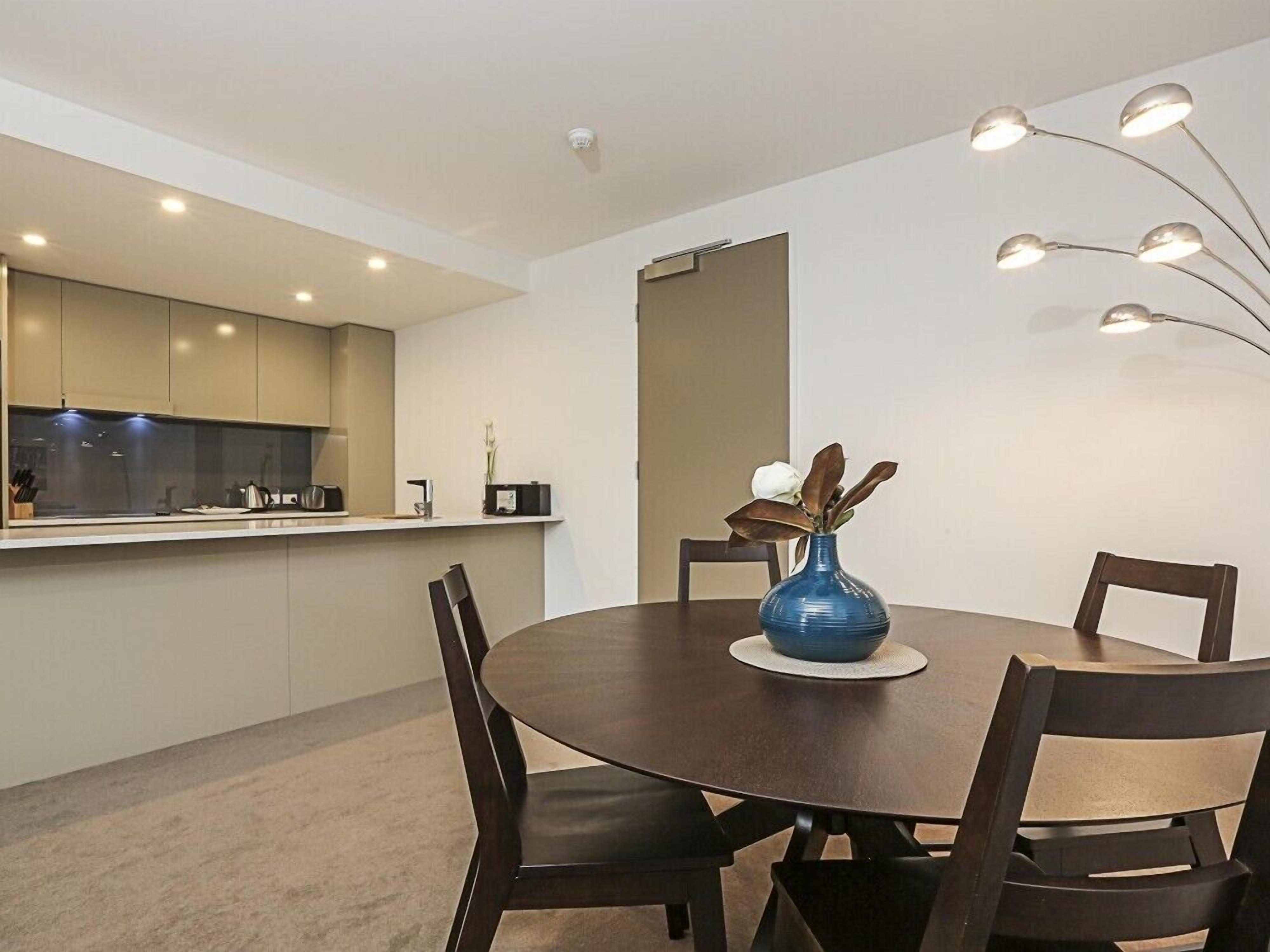 A modern dining room with a round table, four chairs, a vase with flowers, and a kitchen in the background.
