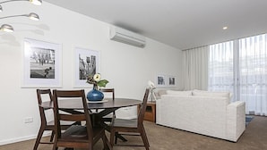 Una sala de estar moderna con comedor, que incluye una mesa redonda con sillas, un sofá y una mesa de centro. Se observan cuadros en las paredes, un equipo de aire acondicionado y un ventanal con cortinas.