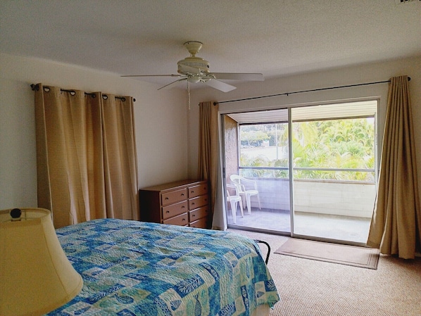 A bedroom with a bed, a ceiling fan, a dresser, and a sliding glass door leading to a balcony with chairs.