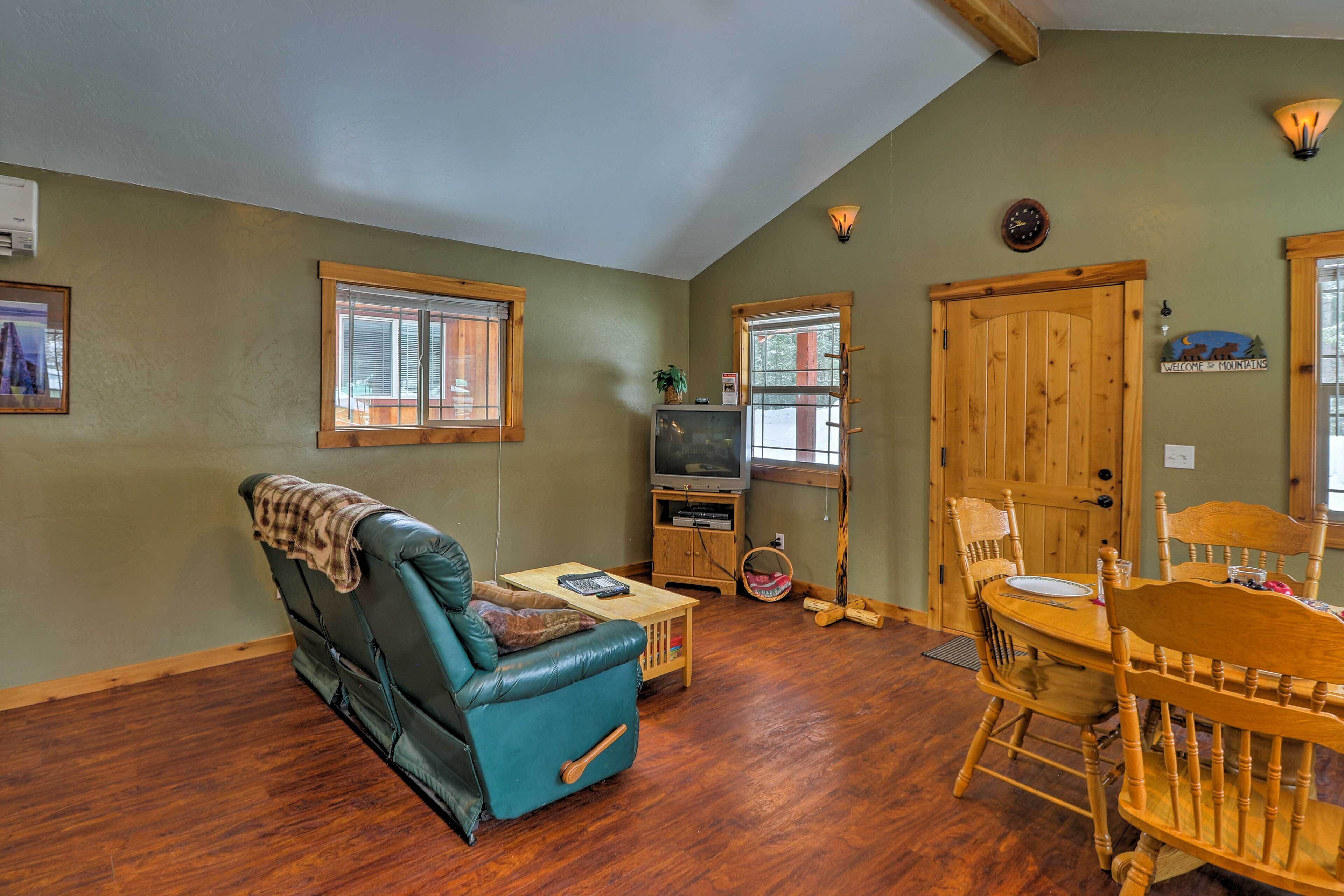 Ein Wohnzimmer mit Ledersessel, Esstisch aus Holz und Stühlen, einem Fernseher und einer Tür mit dem Schild 'Welcome to Mountains'.
