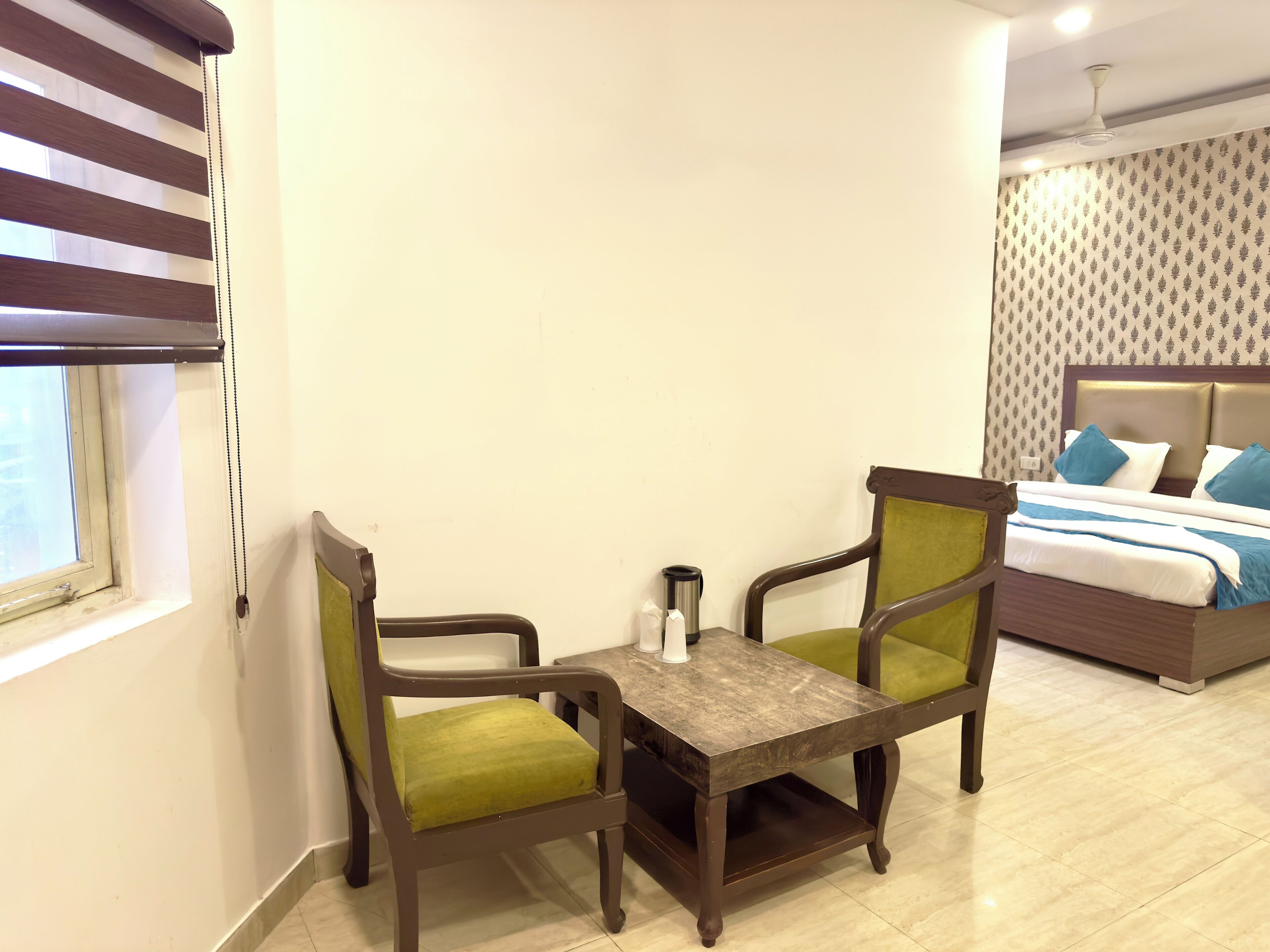 A hotel room with a bed, a small table with chairs, a window with blinds, and a ceiling fan.