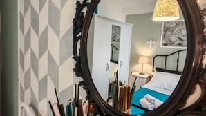 Ein Zimmer mit einer Wand im geometrischen Muster, einem Bett mit blauer Bettdecke, einem weißen Schrank und einem Spiegel mit aufwendiger Rahmung.