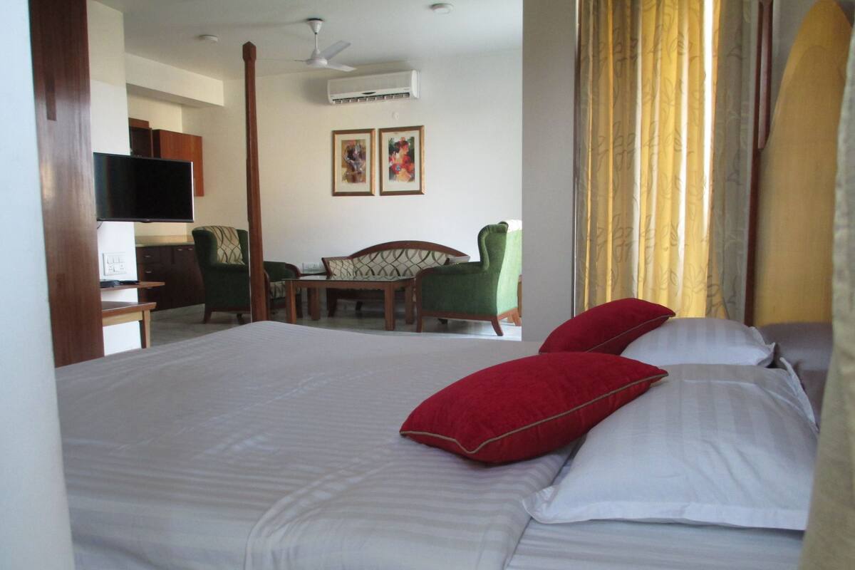 A hotel room with a large bed, a desk with a flat-screen TV, a chair, a table, and two framed pictures on the wall.