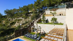 Un espace piscine aménagé sur le toit, avec des chaises longues, une petite piscine et une vue sur une colline verdoyante, parsemée d’arbres et surmontée d’un bâtiment.