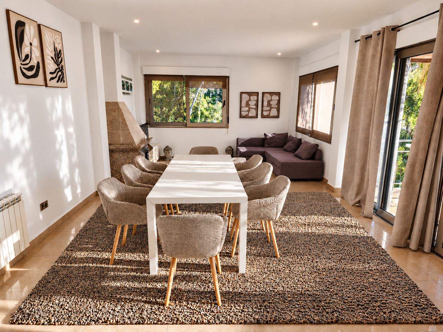 Een moderne woonkamer met een eethoek, met een lange tafel, rieten stoelen, een bank en een kamerkleed met een patroon.