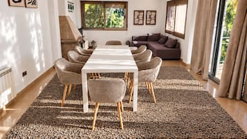 Een moderne woonkamer met een eethoek, met een lange tafel, rieten stoelen, een bank en een kamerkleed met een patroon.