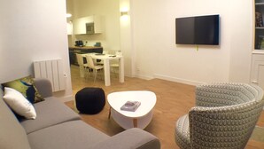 Una sala de estar con un sofá gris, una mesa de centro redonda y un sillón estampado. En el fondo, se ve un comedor con una mesa y sillas blancas.