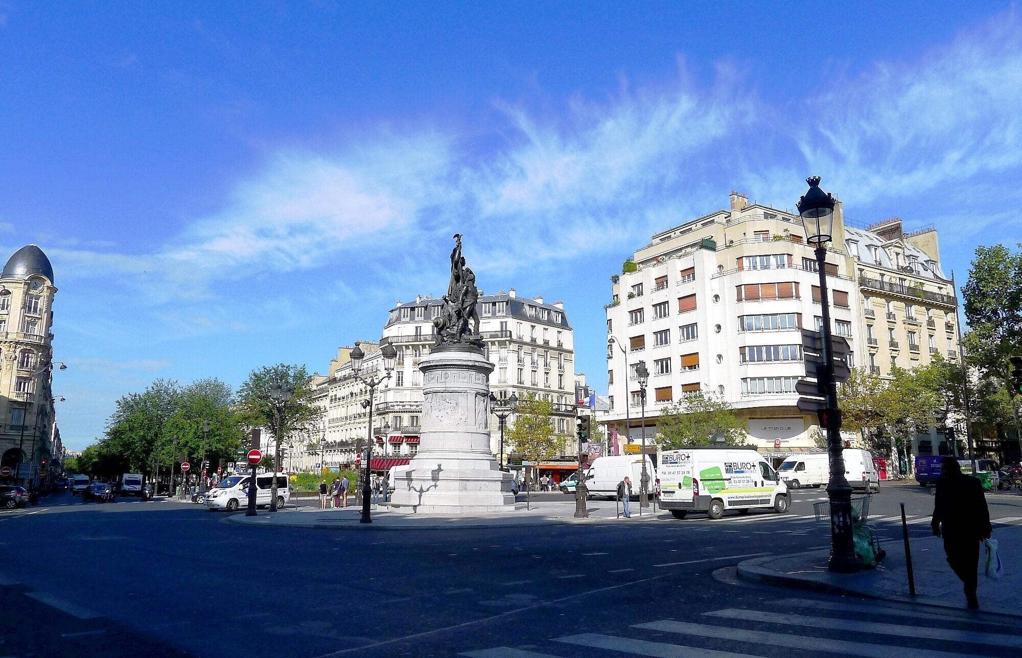 A city square with a central monument, surrounded by multi-story buildings with balconies.