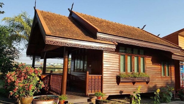 A wooden house with a sloped roof, a porch with decorative railings, and a window with a plant box. - Hugphaeng Garden (Ban Phaeng)