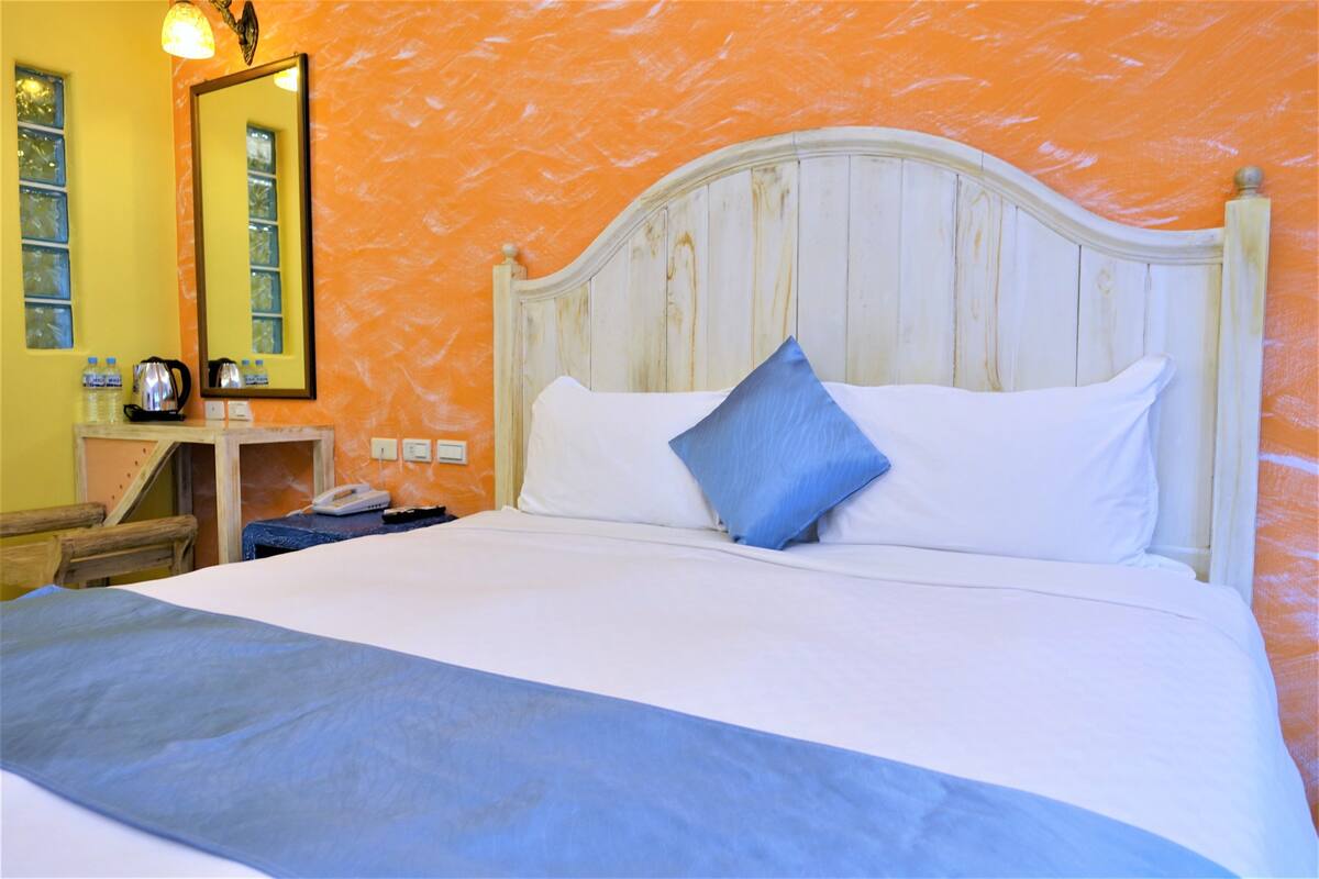 Ein Bett mit weißer Bettwäsche und einem blauen Kopfkissen, ein Nachttisch mit einem Wasserkocher und ein Spiegel an der Wand.
