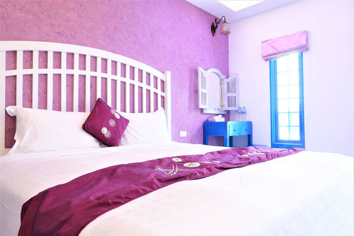 Ein Bett mit weißem Kopfteil, einer violetten Tagesdecke und einem einzelnen violetten Kissen.