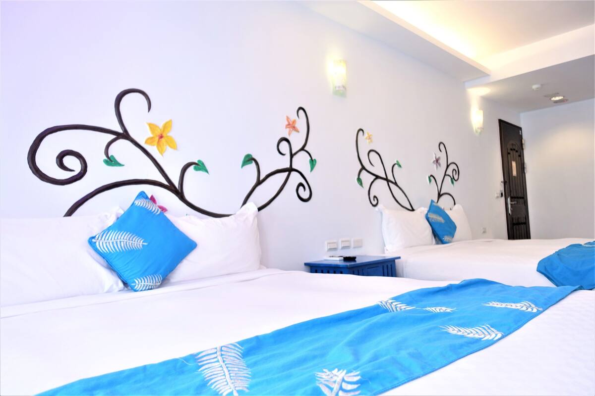 Ein Hotelzimmer mit zwei Betten, weißer Bettwäsche, blauen Zudecken und Wandkunst. Ein Nachttisch mit Lampe ist ebenfalls zu sehen.