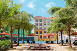 A hotel with a pool area, palm trees, and red umbrellas.