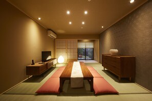 A traditional Japanese room with tatami flooring, a low table with a table runner, a wooden dresser, a television on a side table, and sliding doors leading to a garden.