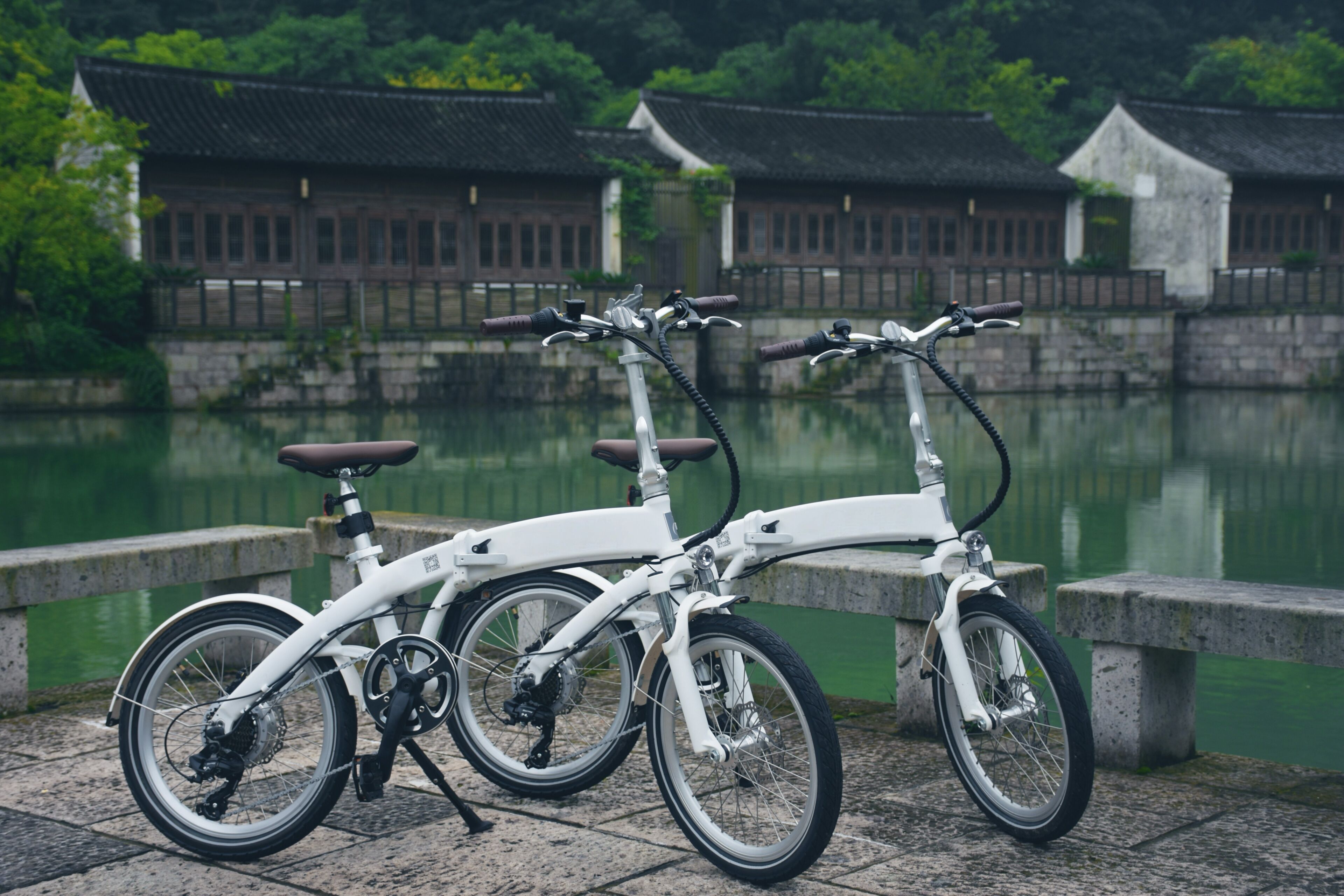 Two white bicycles parked by a stone wall with a body of water and traditional buildings in the background.