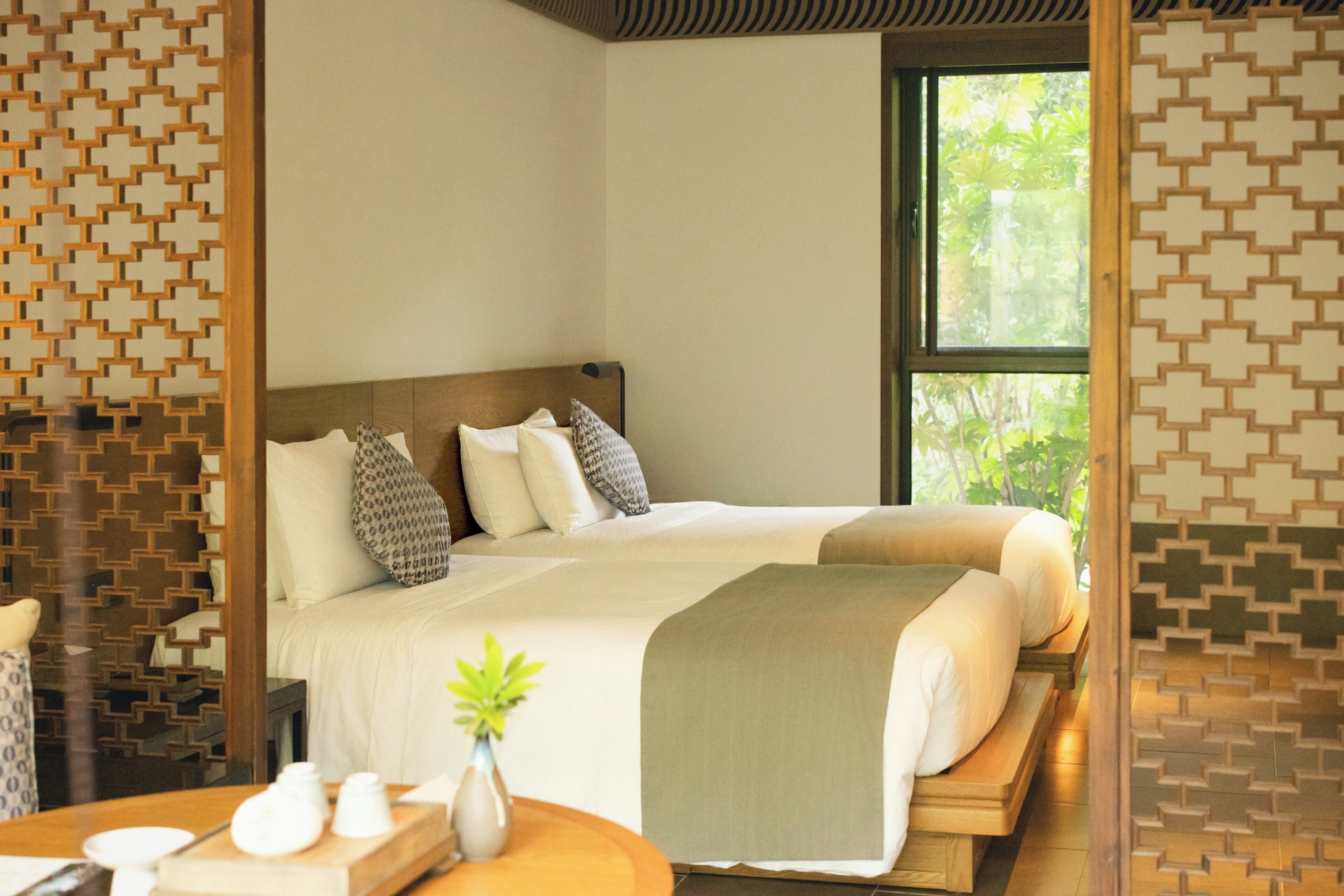A room with a large bed, a bedside table with a vase, and a window with a view of greenery.