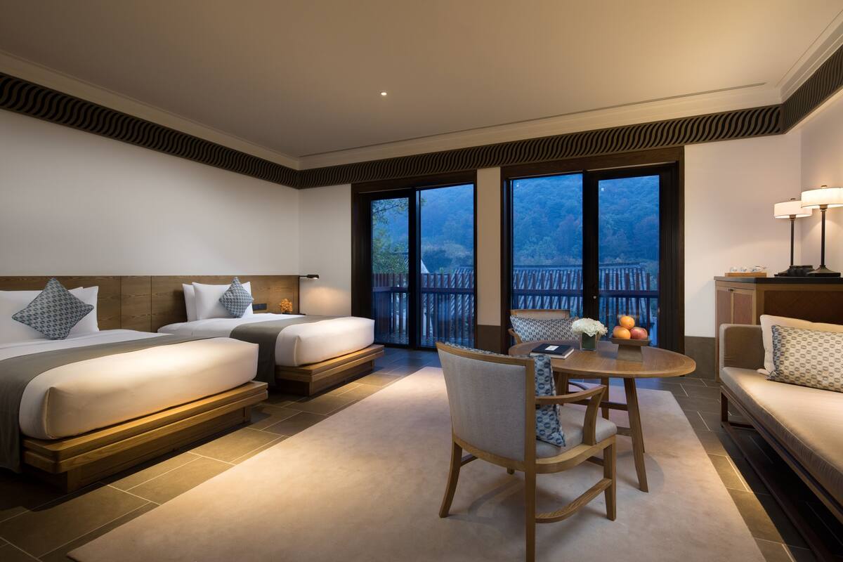 A hotel room with two beds, a sofa, a dining table, and a balcony with a view.