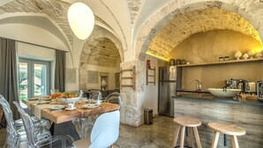 A rustic kitchen with stone arches, a wooden dining table with chairs, a refrigerator, and a sink.