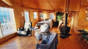 A cozy cabin interior with a wooden interior, a bed, a seating area, a fireplace, and a kitchen area. - Cliff Waters Cottage, Private oceanfront secluded relaxation, Pleasant Bay, NS (Pleasant Bay)