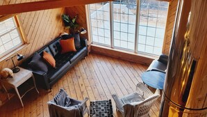A cozy cabin interior with a black leather sofa, a small table, wicker chairs, and a large window offering a view of the outdoors. - Cliff Waters Cottage, Private oceanfront secluded relaxation, Pleasant Bay, NS (Pleasant Bay)