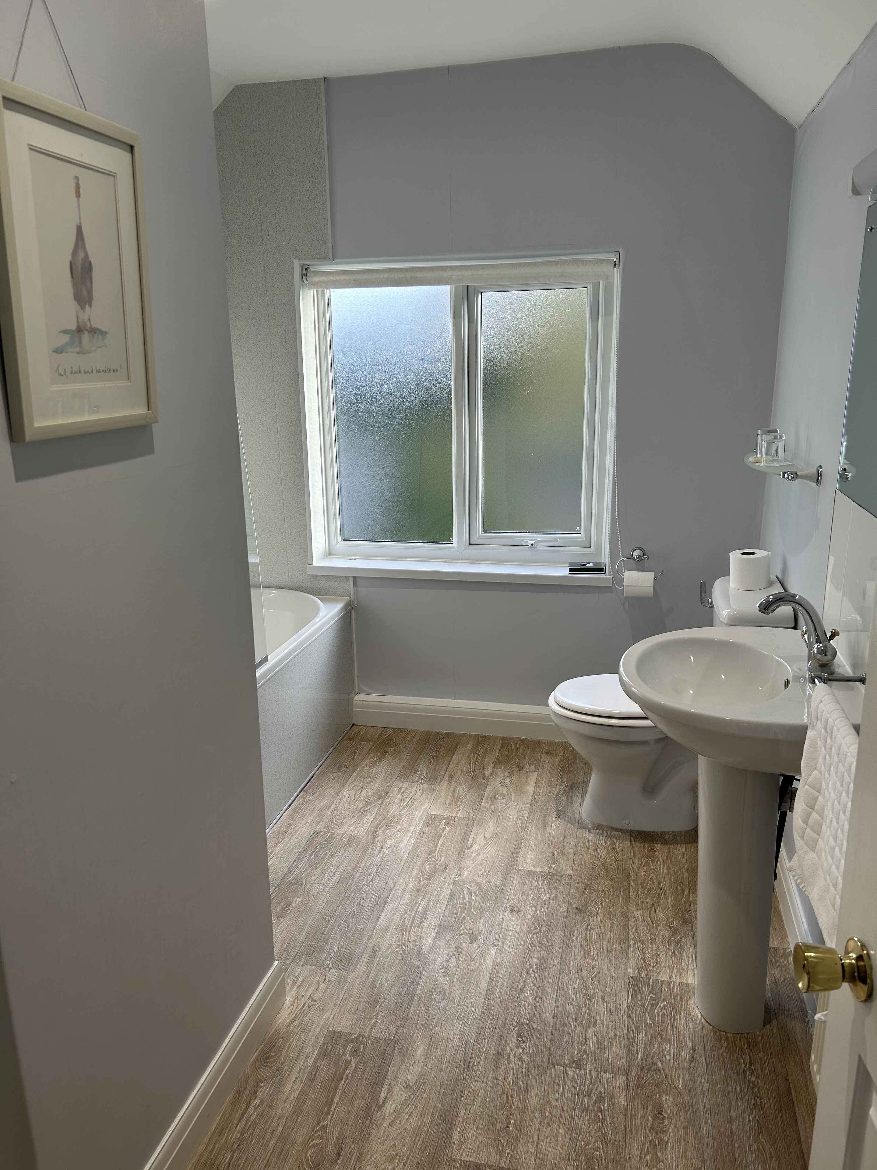 Une salle de bain équipée d’une baignoire, de toilettes et d’un lavabo, avec un plancher en bois et une fenêtre en verre dépoli.