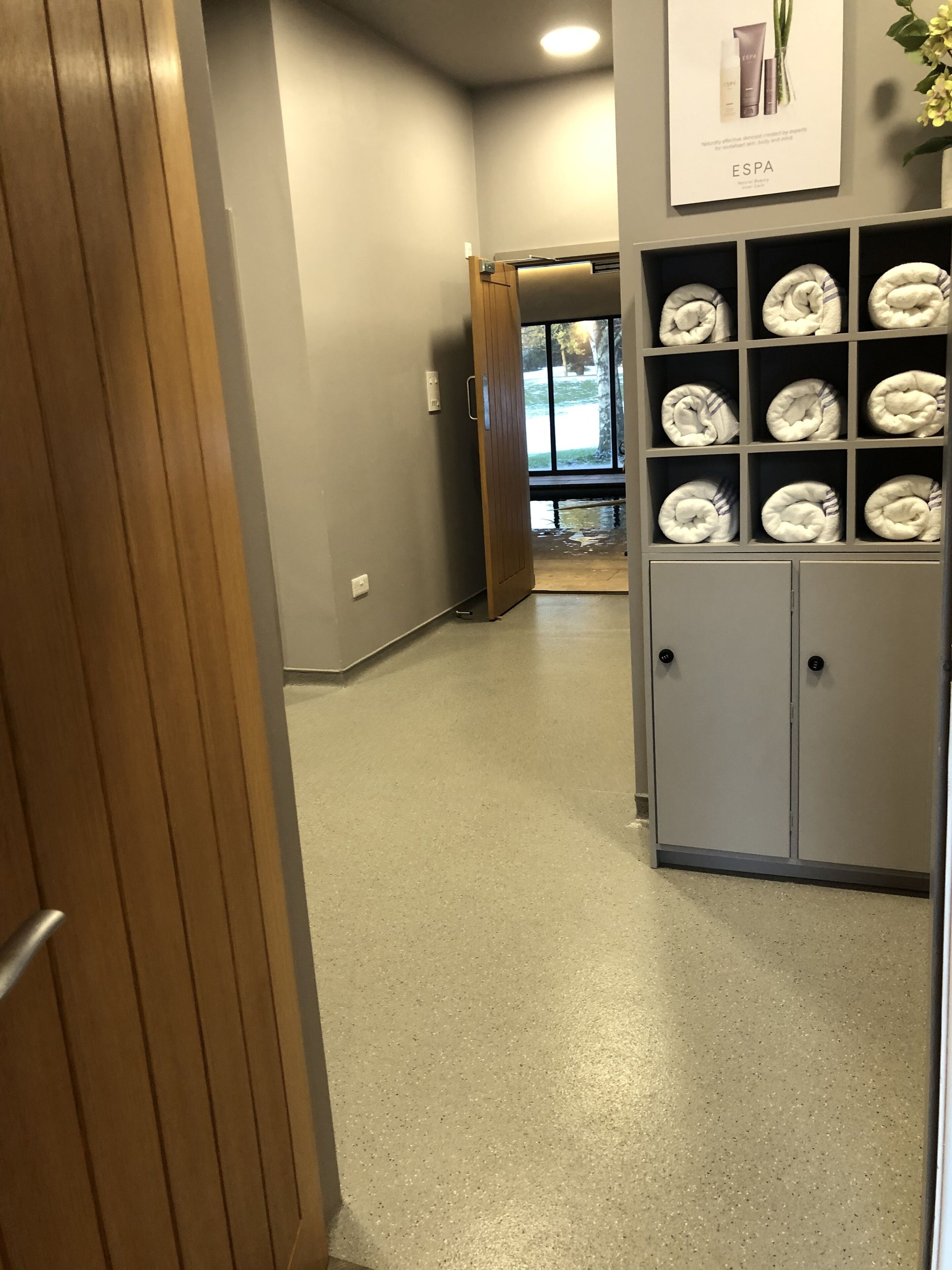 A hallway with a wooden door, a wall-mounted shelf with folded towels, and a door leading to a room with a view of trees.