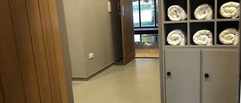 A hallway with a wooden door, a wall-mounted shelf with folded towels, and a door leading to a room with a view of trees.