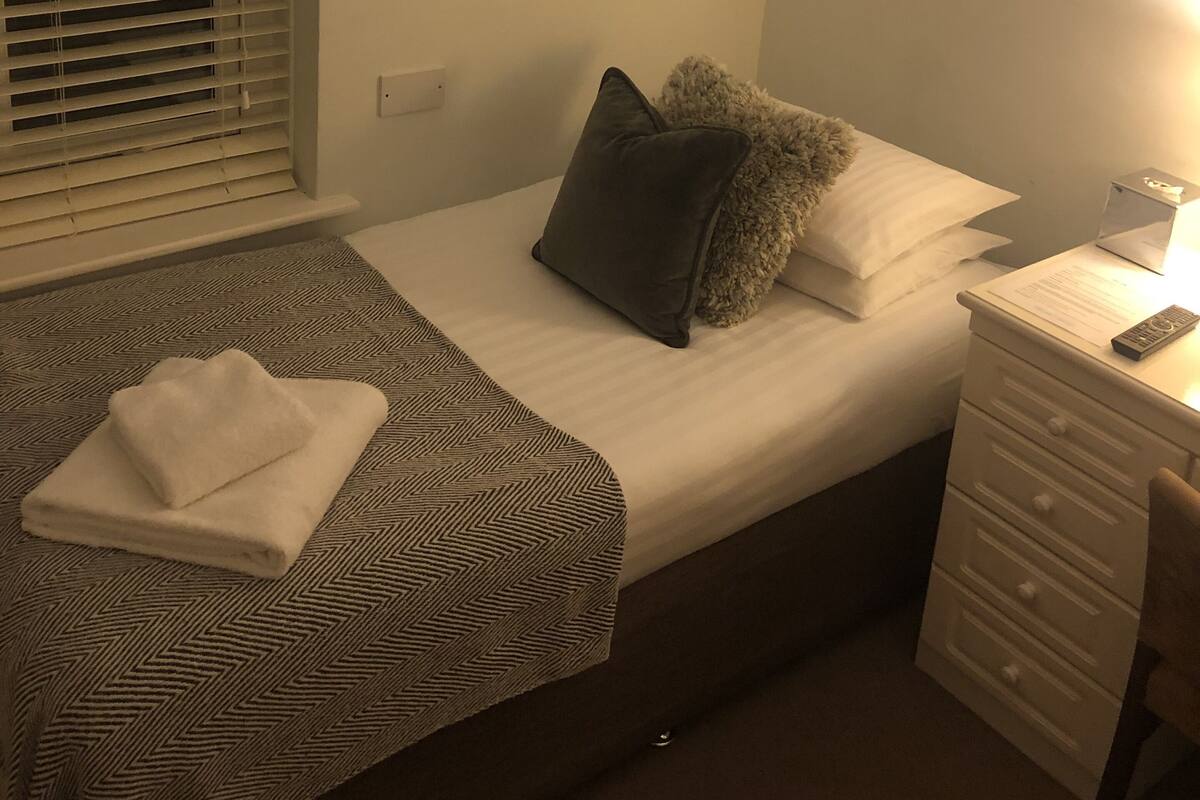 A bedroom with a bed, a small desk, a chair, a window with blinds, and a wall-mounted picture.