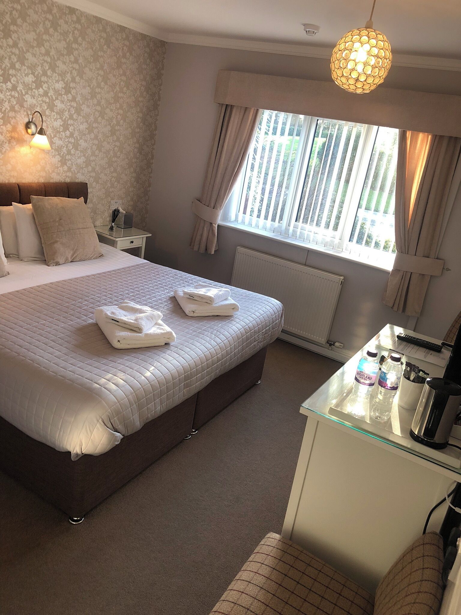A neatly arranged bedroom with a large bed, bedside tables, a desk with a kettle, and a window with curtains.