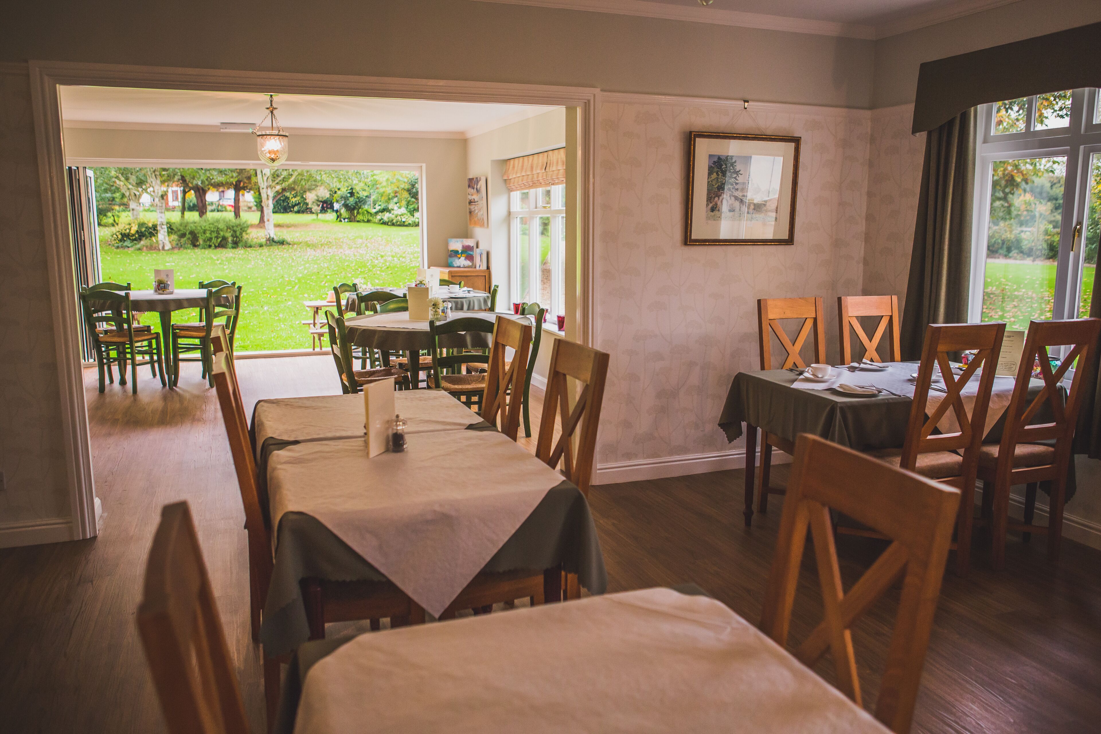 A dining area with tables and chairs, a view of the garden, and a framed picture on the wall.