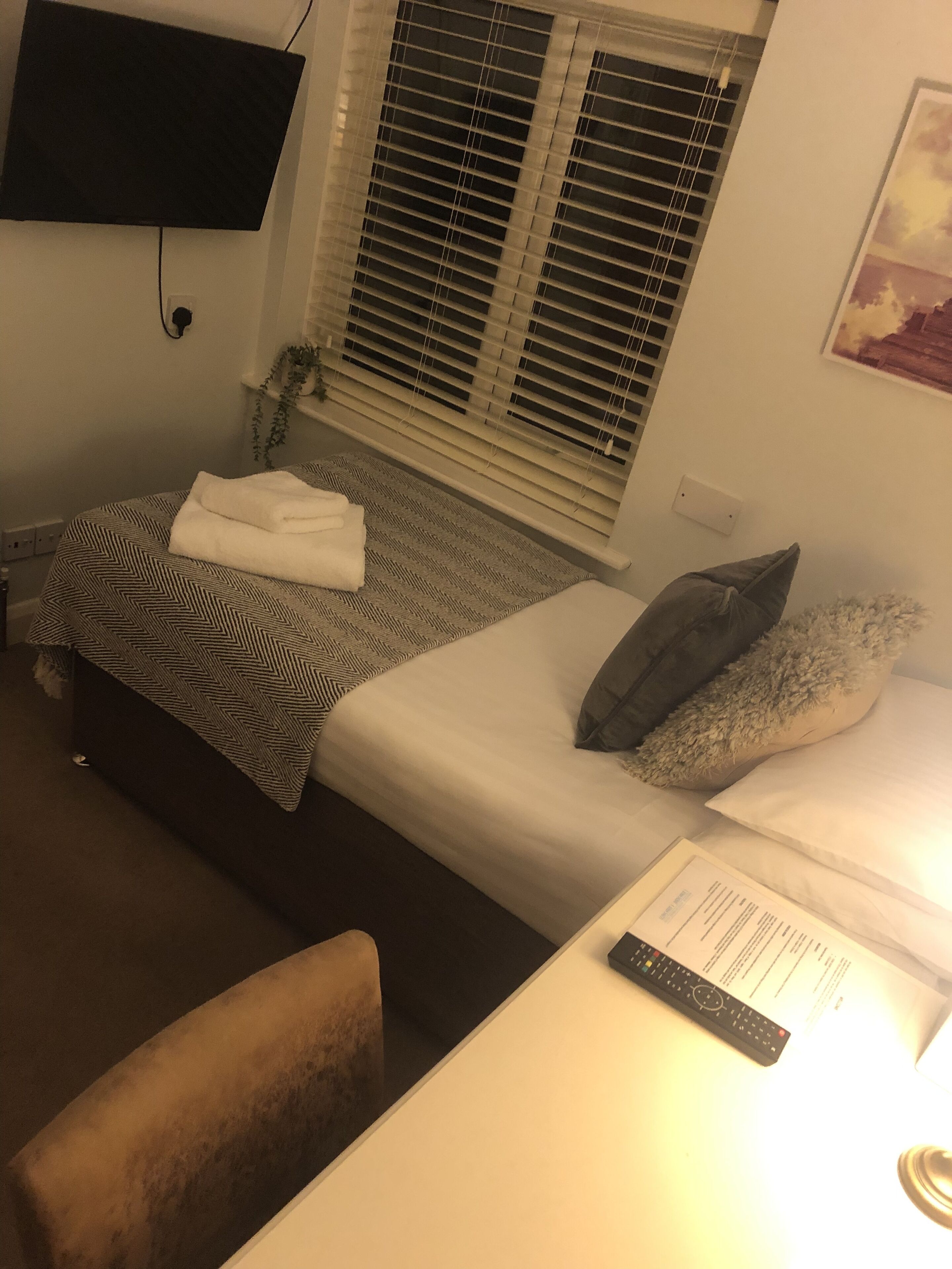 Ein kleiner, schwach beleuchteter Raum mit zwei Betten, einem Schreibtisch und einem an der Wand befestigten Fernseher.