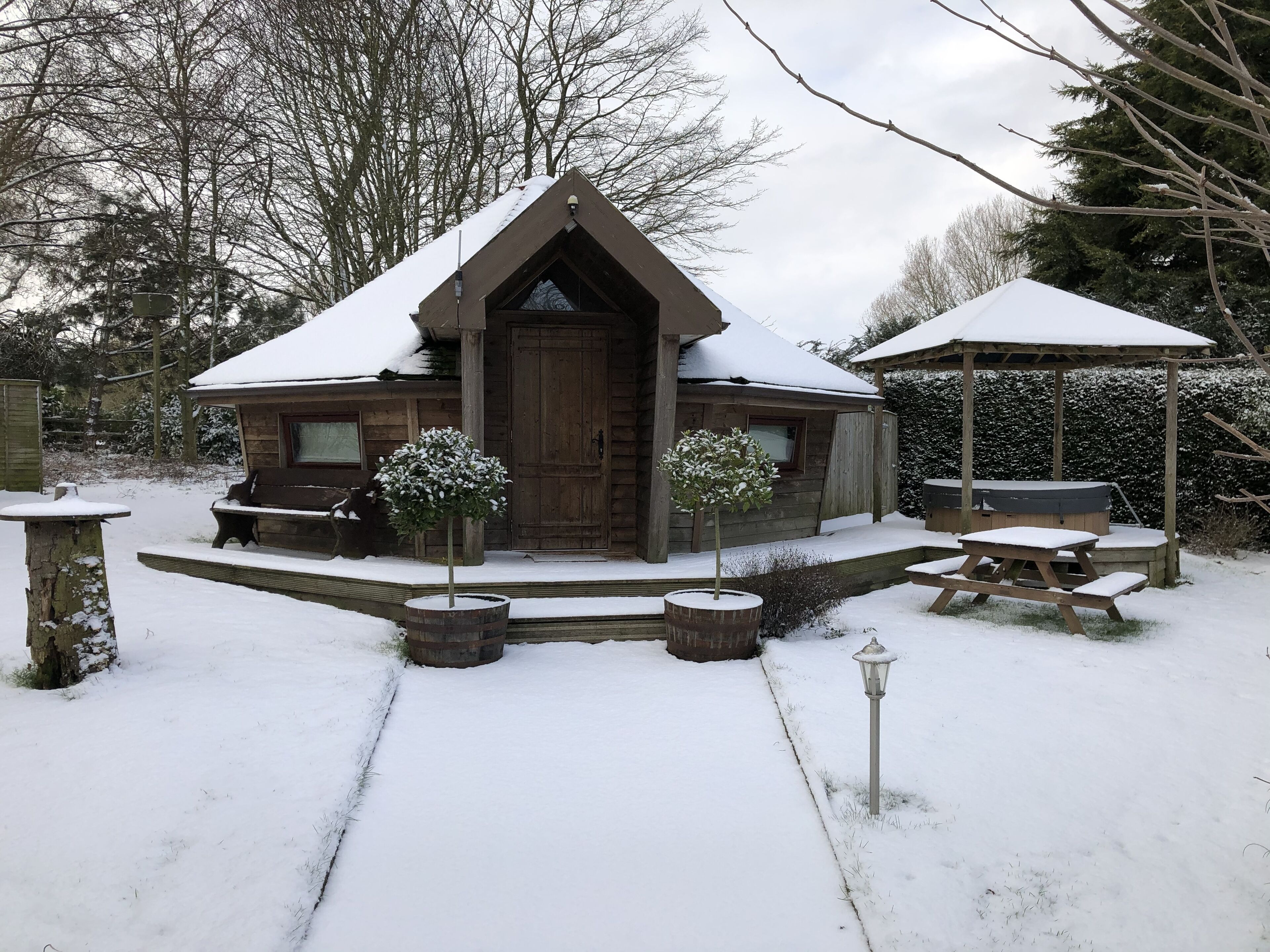 A wooden cabin with a snow-covered roof, a porch with benches, and a hot tub.