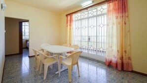 Una habitación con una mesa de comedor y sillas, un ventanal con celosía metálica y una puerta que da a otra estancia.
