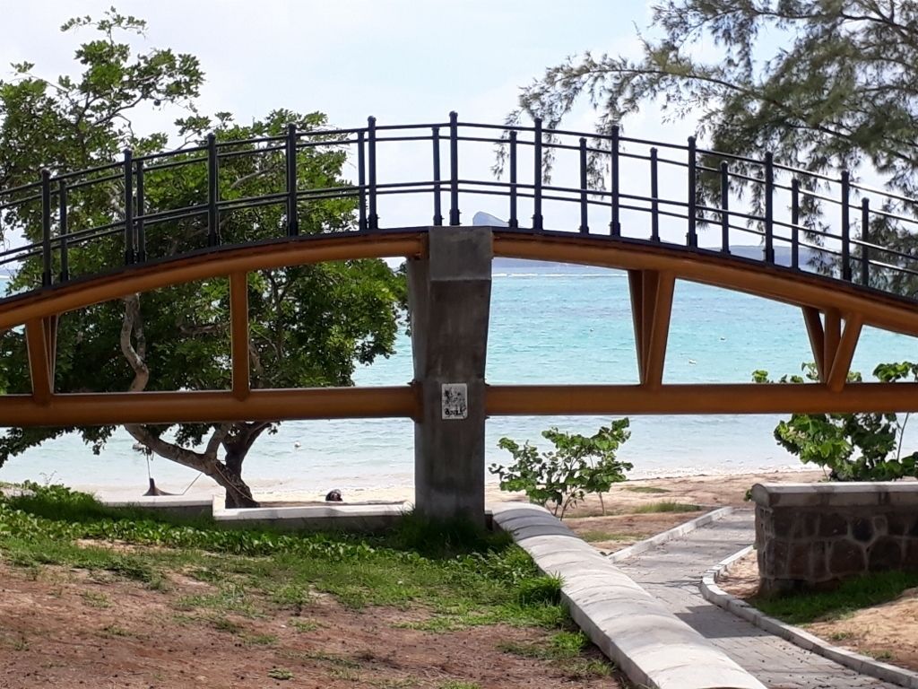 A pedestrian bridge with a metal railing and a concrete base, overlooking a beach and the sea.