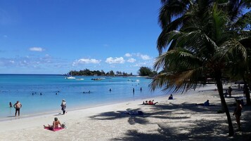 Una playa con agua azul cristalina, gente nadando y tomando el sol, y barcos a lo lejos.