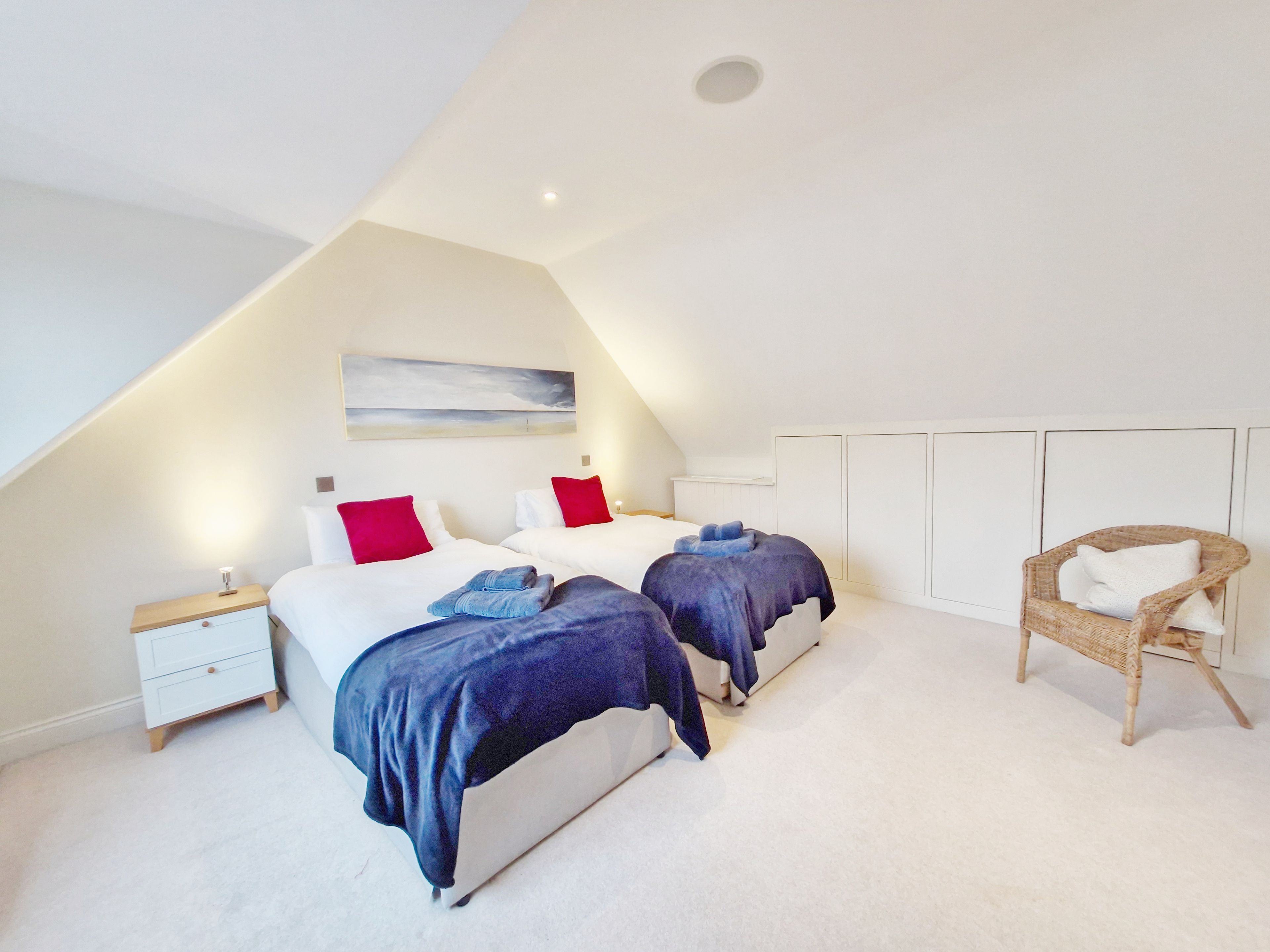A bedroom with two beds, a small wooden nightstand, a wicker chair, and a white ceiling.