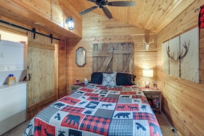 Une chambre dans une cabane en bois, comprenant un lit, une commode, une table de chevet, un ventilateur de plafond et des œuvres d’art accrochées au mur.