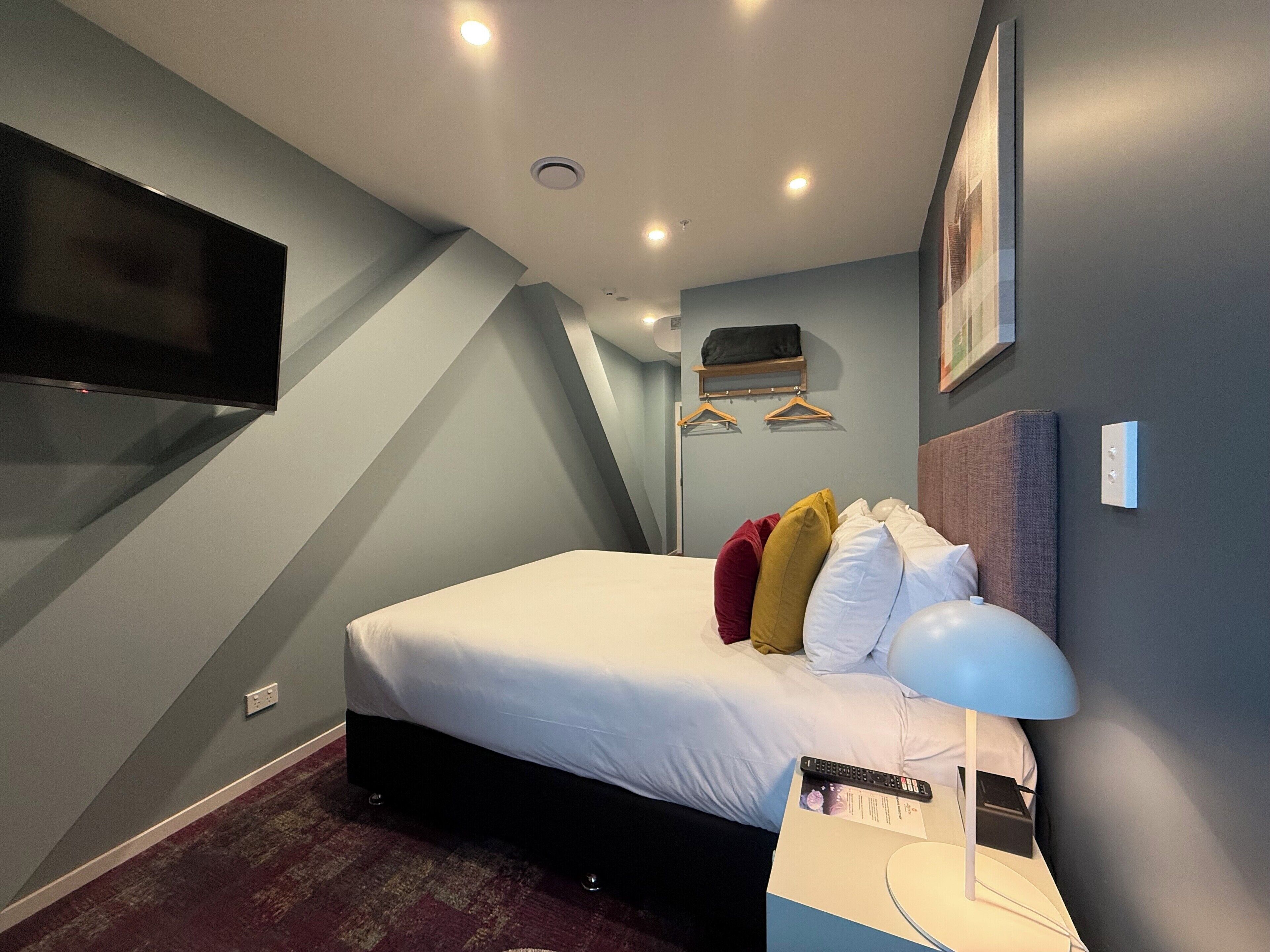 Une chambre d’hôtel moderne avec un grand lit, un téléviseur à écran plat fixé au mur, une table de chevet avec une lampe et un tableau encadré au-dessus du lit.