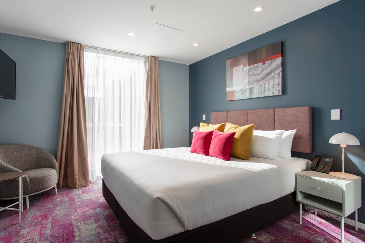 A hotel room with a large bed, a chair, a nightstand, and a window with curtains.